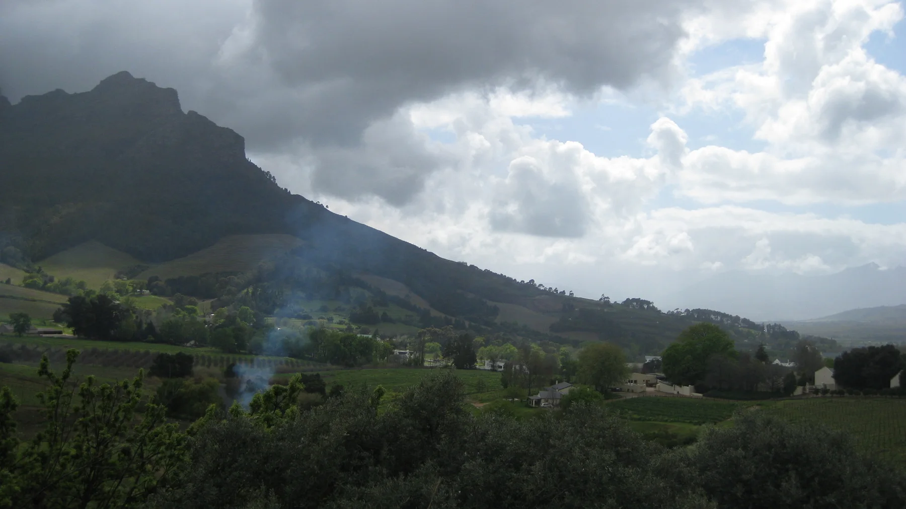 Green fields and trees occupy a valley beneath steep mountain slopes and broken clouds.
