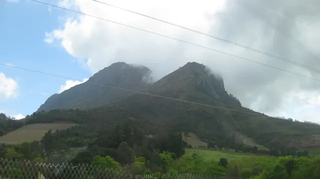 Two steep mountain peaks rise beyond a roadside fence and green slopes, with clouds near their tops.