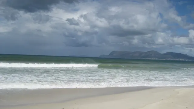 White waves break along the beach beneath dark clouds and a distant mountain coastline.