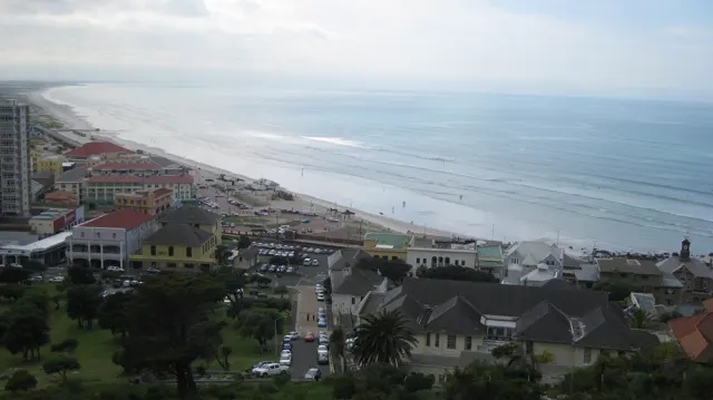 An elevated view follows roads and buildings toward a long beach and pale blue sea.