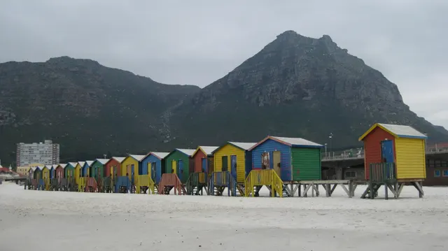 Brightly painted beach huts stand in a row on the sand below a steep mountain.