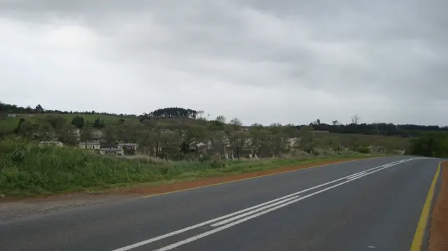 A paved road bends past low vegetation and fenced fields beneath a gray sky.