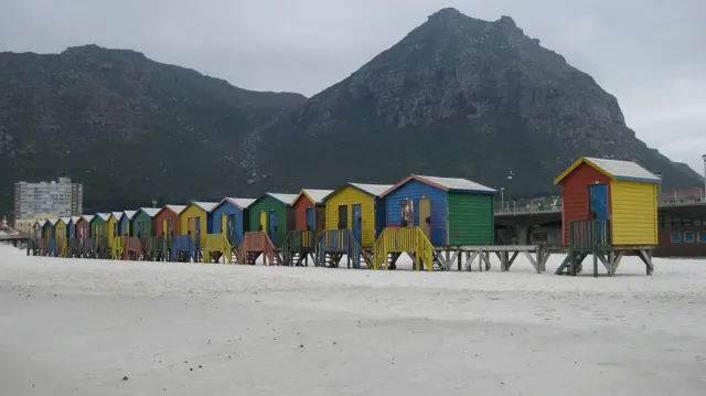 Bright beach huts line pale sand below a dark mountain.