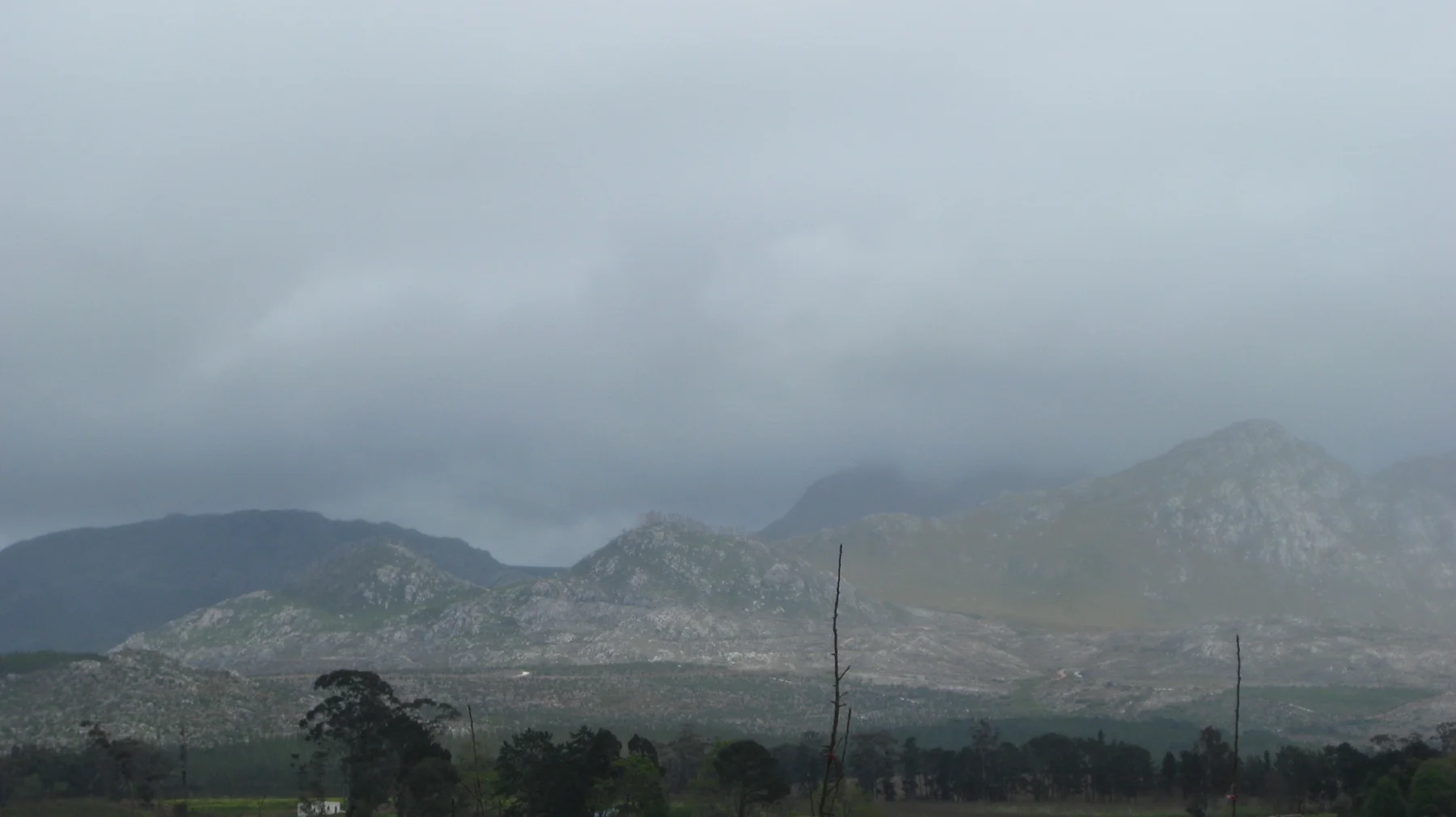 Low cloud veils a rugged mountain range beyond trees and fields.