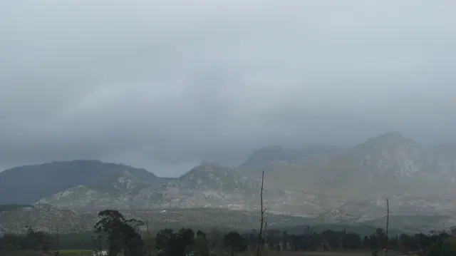 Low cloud veils a rugged mountain range beyond trees and fields.