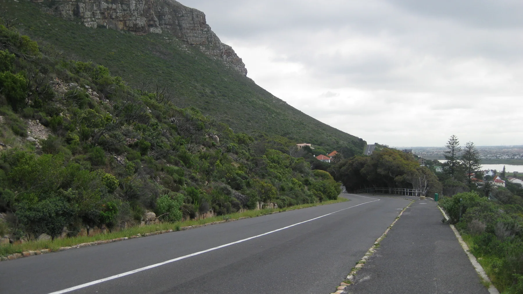 A road descends beside a green rocky slope toward seaside buildings.