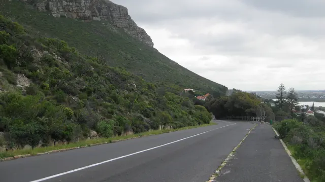 A road descends beside a green rocky slope toward seaside buildings.