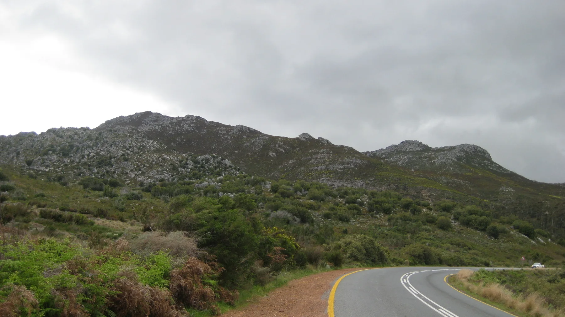 A double-lined road curves through low vegetation beneath rocky hills.
