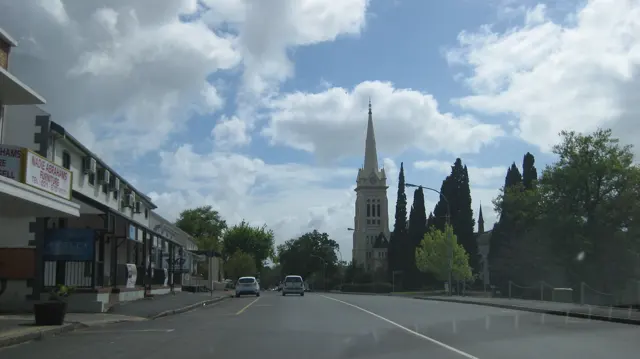 A tall church spire rises ahead of a street beneath bright clouds.
