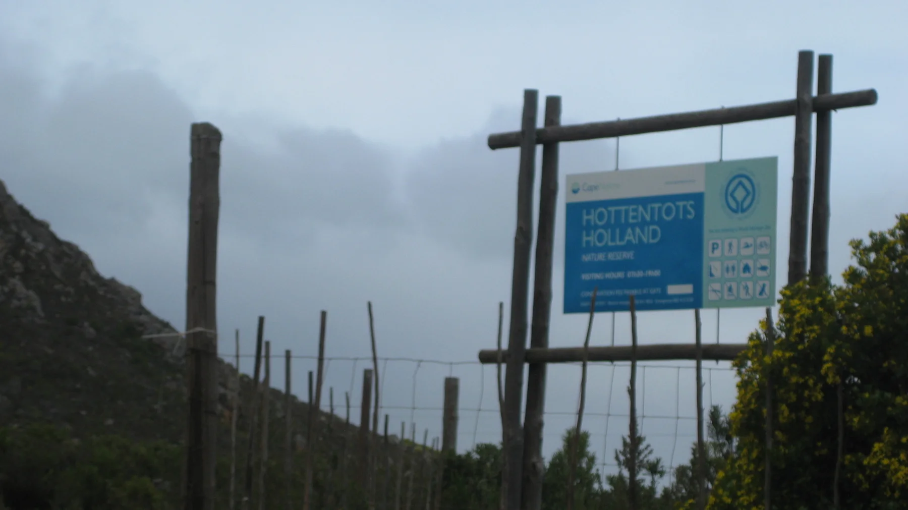 A blue roadside sign stands among wooden posts below cloud-covered mountains.