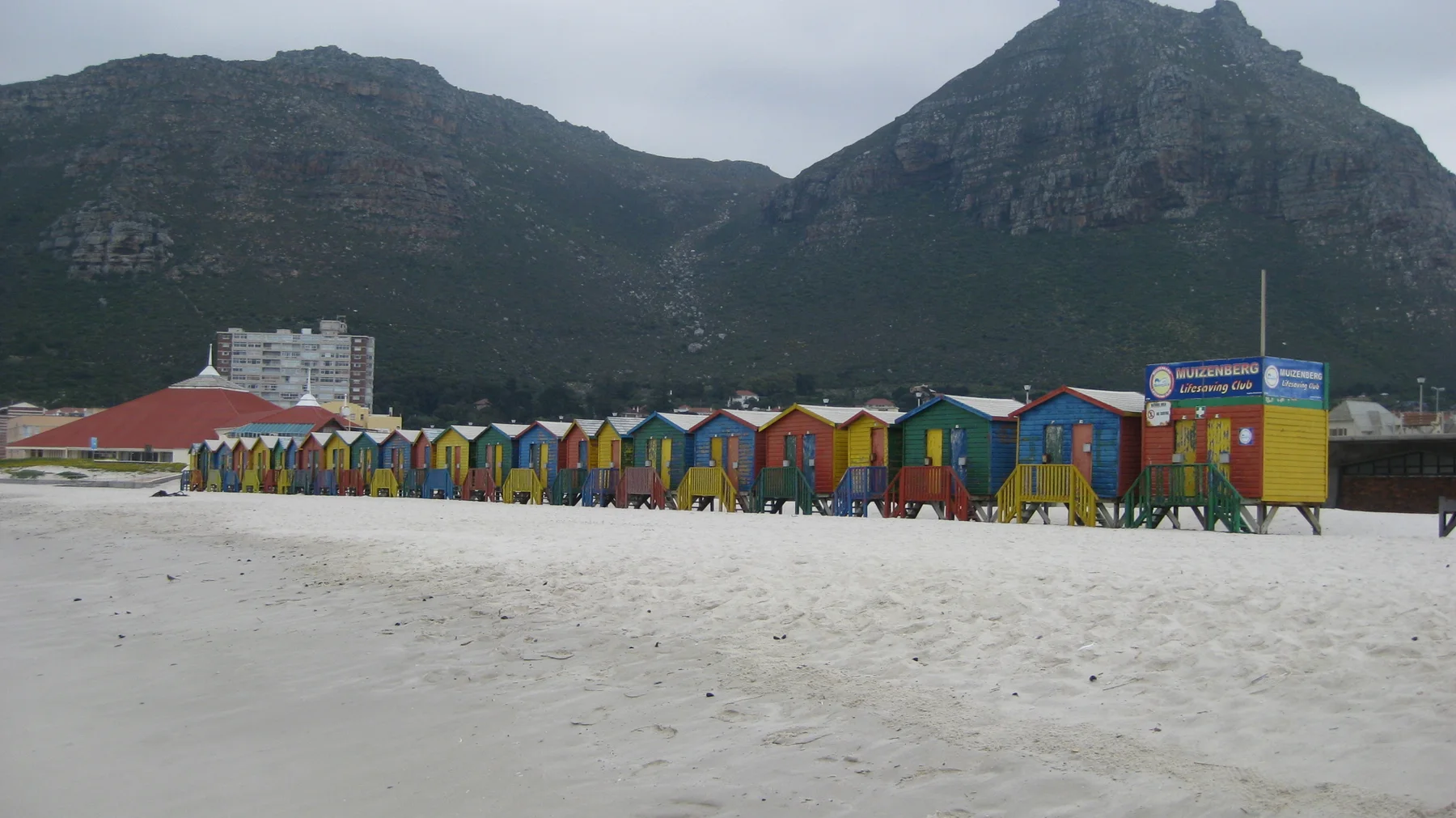 A long row of colorful huts stretches across the beach below a mountain.