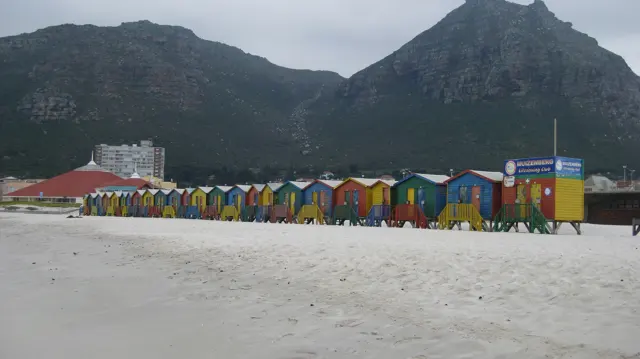 A long row of colorful huts stretches across the beach below a mountain.
