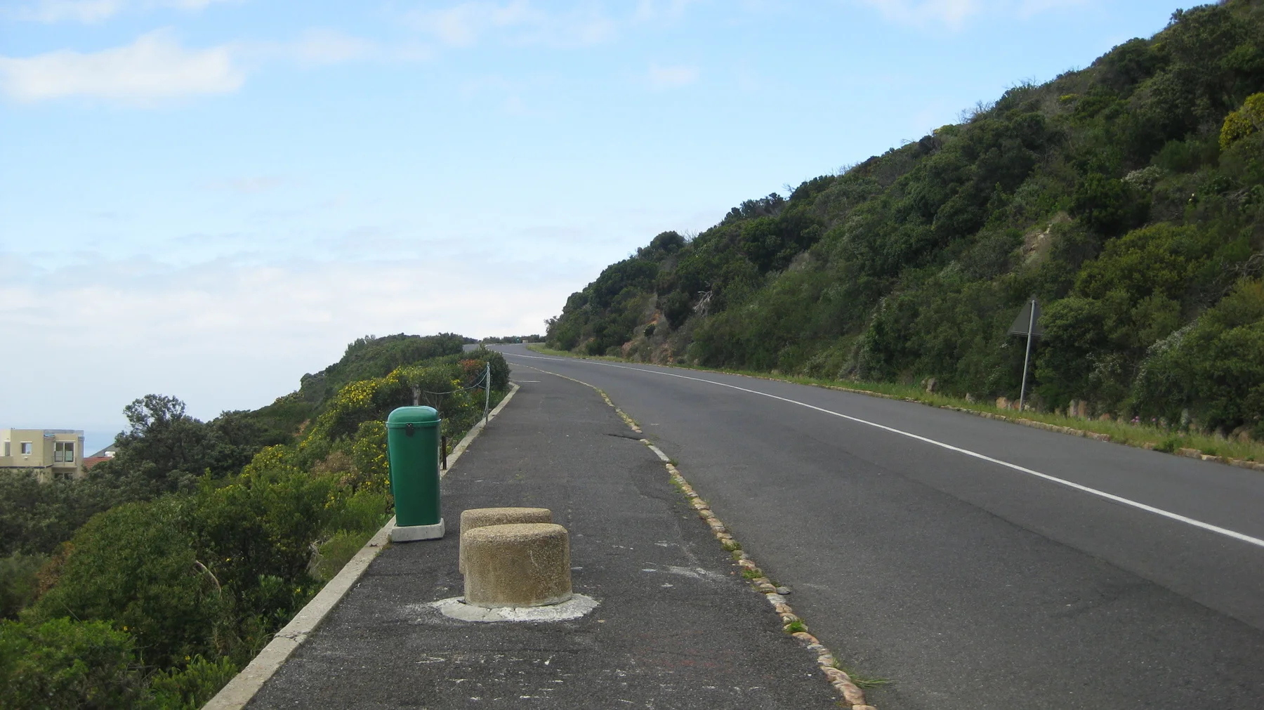 A bin and short concrete post stand beside a coastal mountain road.