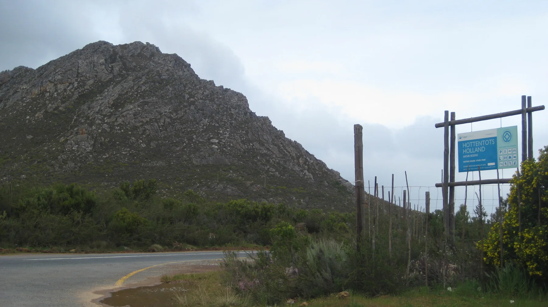 A roadside sign and fence stand beside a bend below a rocky mountain.