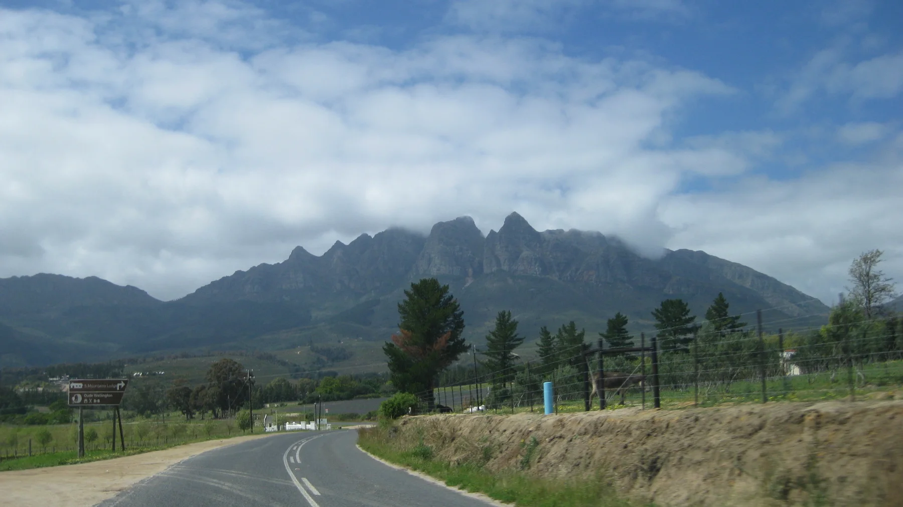 A road leads toward a jagged mountain skyline past fields and trees.