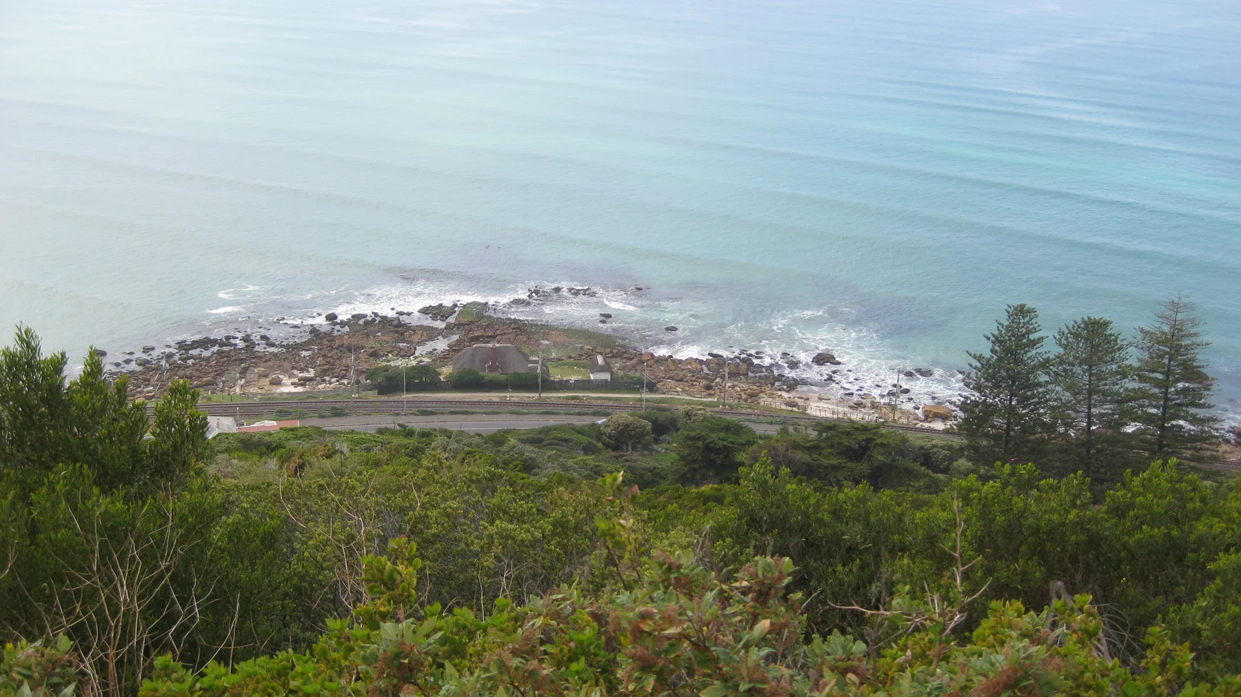 A rocky coastal point lies below dense green shrubs and calm water.