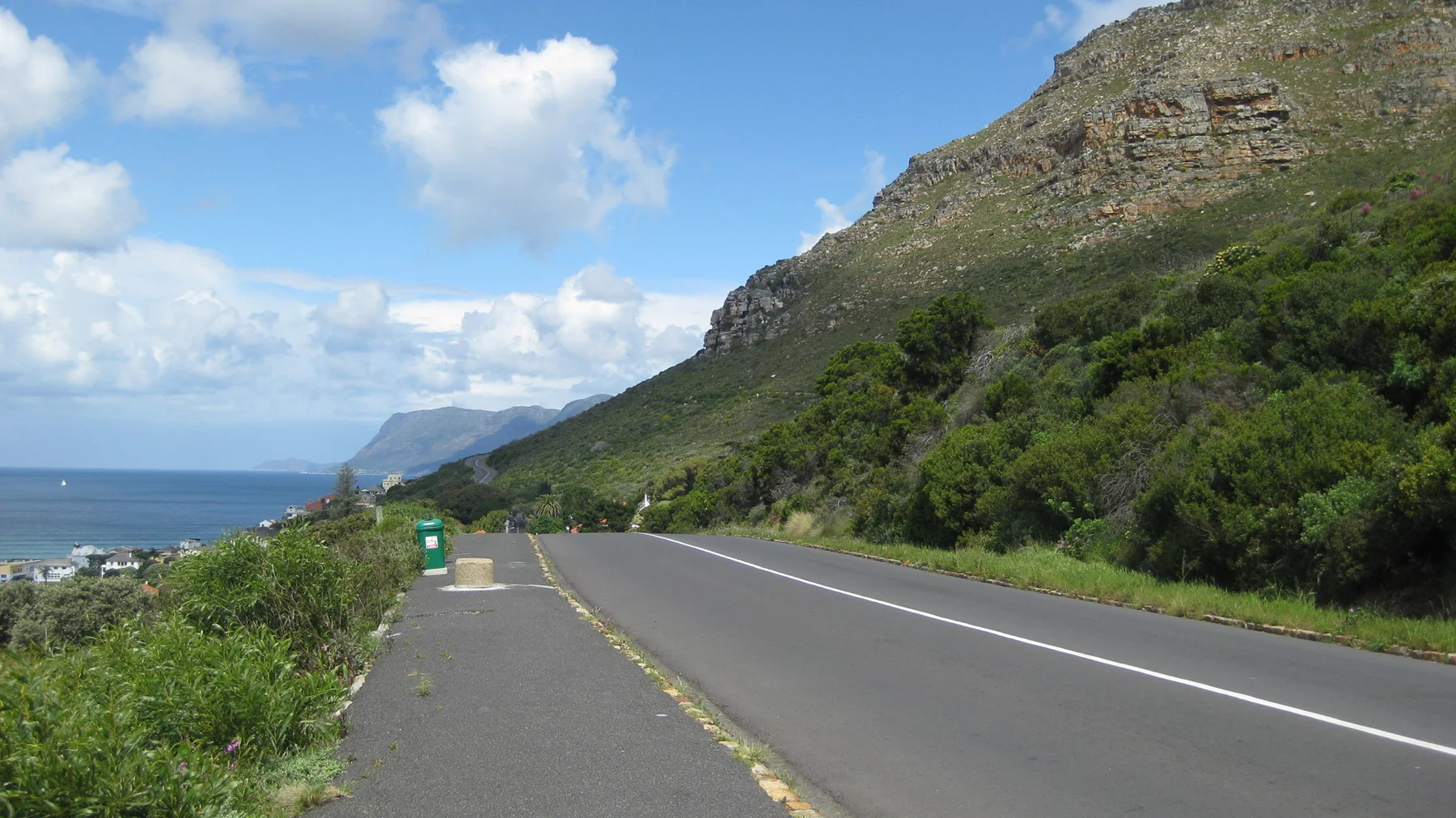 A coastal road runs below a rocky green slope with blue ocean beyond.