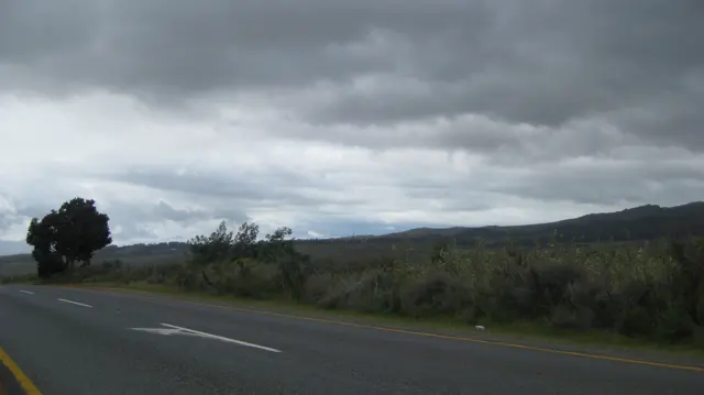 Dark clouds hang above a road bordered by low countryside.
