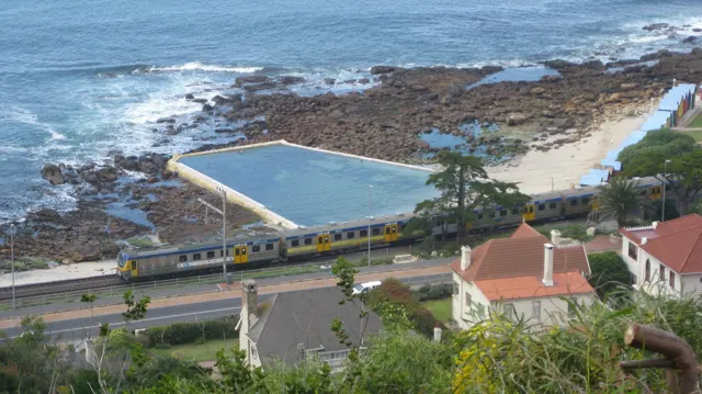 A rectangular coastal pool lies beside rocky shore, railway tracks and buildings.