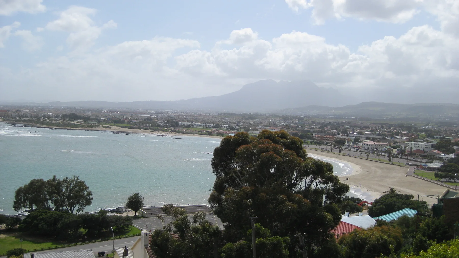 A long beach curves around blue water below a hazy town and mountain backdrop.