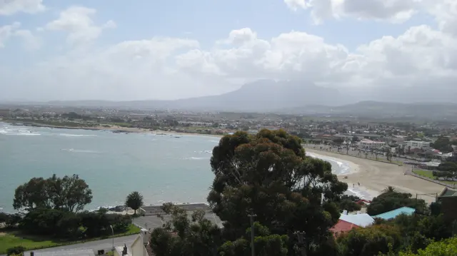 A long beach curves around blue water below a hazy town and mountain backdrop.