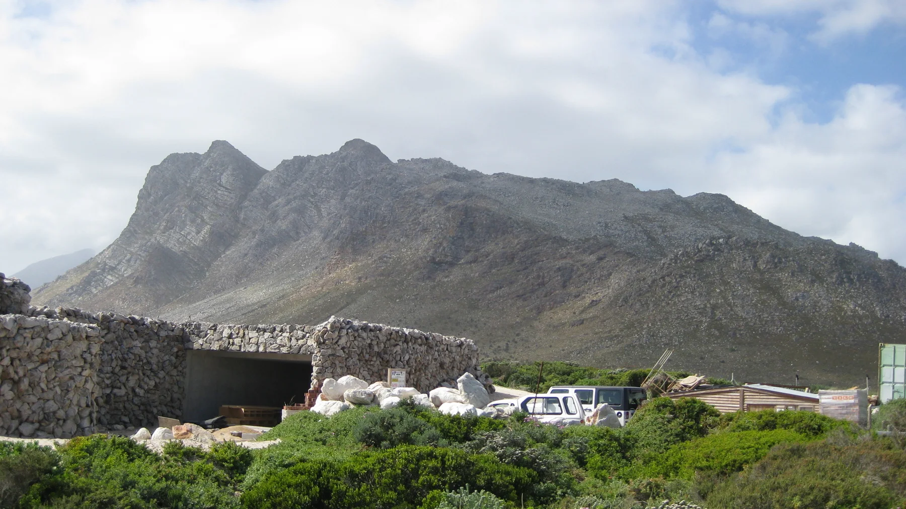 A low stone structure and parked vehicles sit below a rugged mountain ridge.
