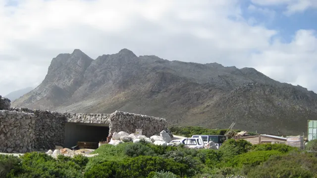 A low stone structure and parked vehicles sit below a rugged mountain ridge.