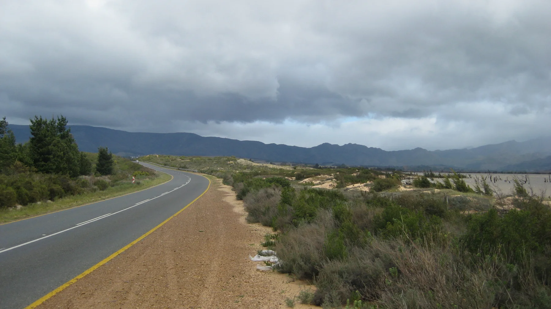 A quiet road follows scrubland and water toward dark distant hills.