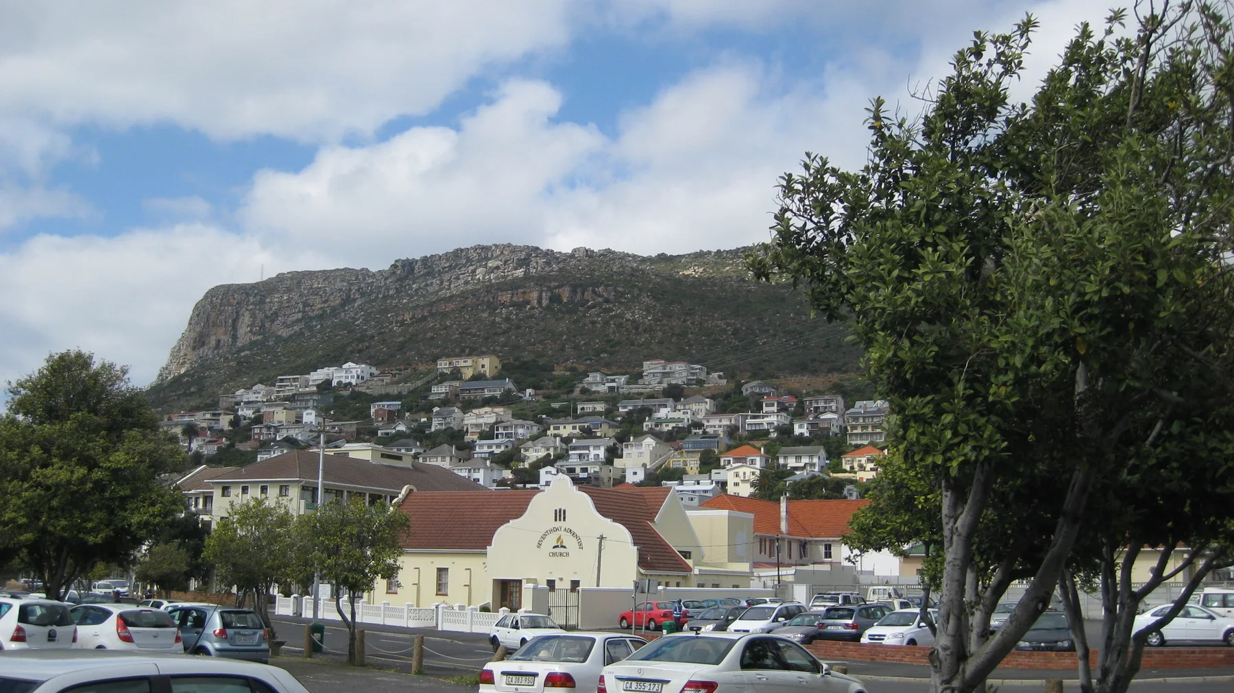 A pale gabled building stands beneath hillside homes and a rocky ridge.