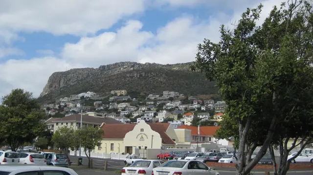 A pale gabled building stands beneath hillside homes and a rocky ridge.