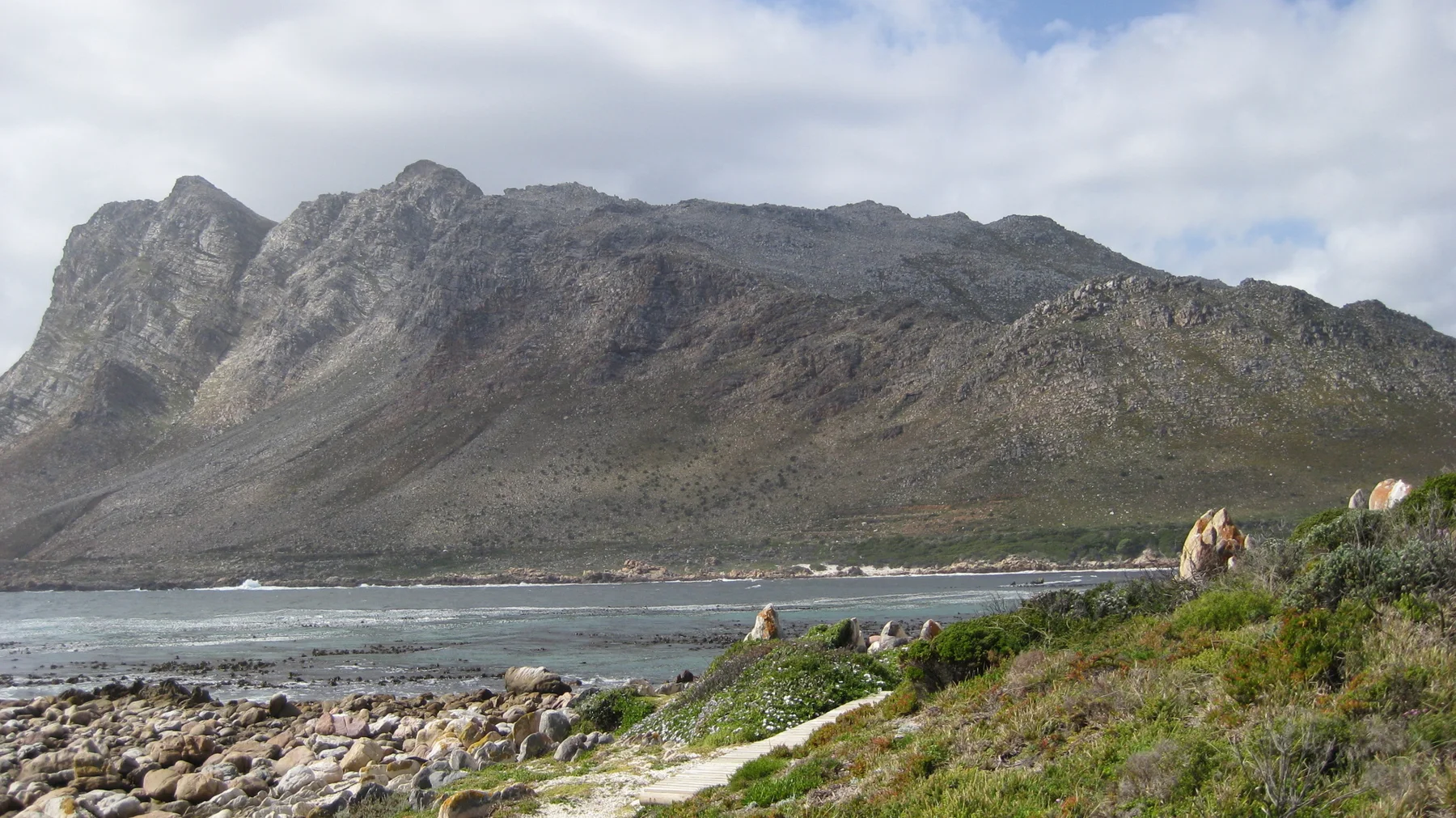 A long rocky mountain rises across water beyond a stony shoreline.