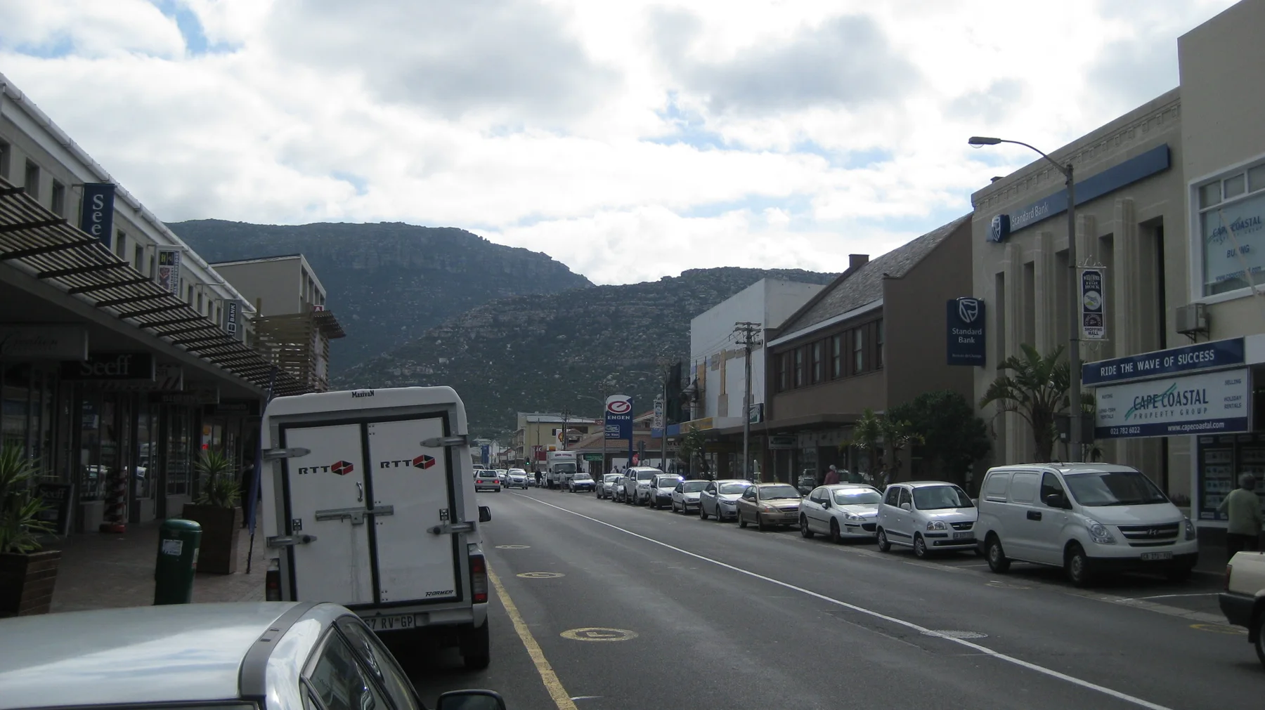 Parked vehicles and low buildings line a street toward the mountains.