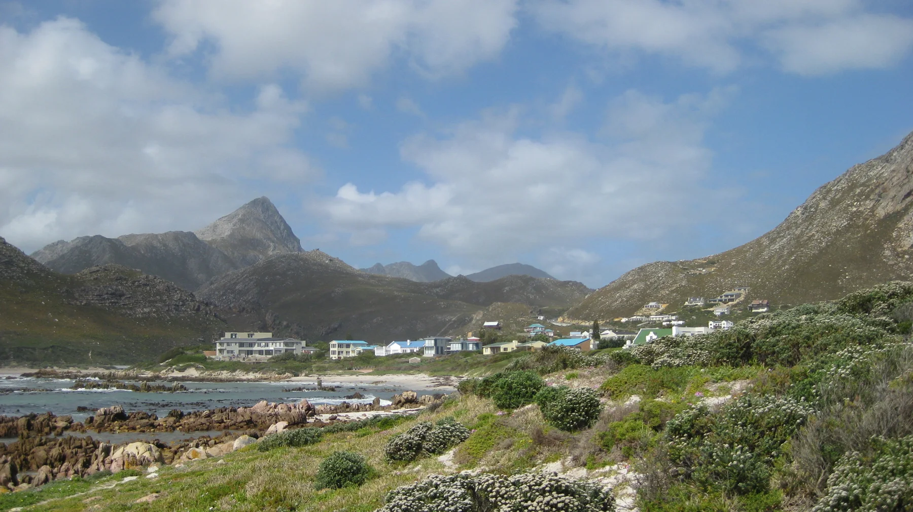 Pale coastal buildings sit between a rocky shore and mountain slopes.