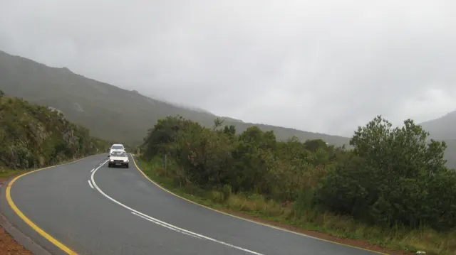 A white vehicle approaches along a curving road beneath misty hills.