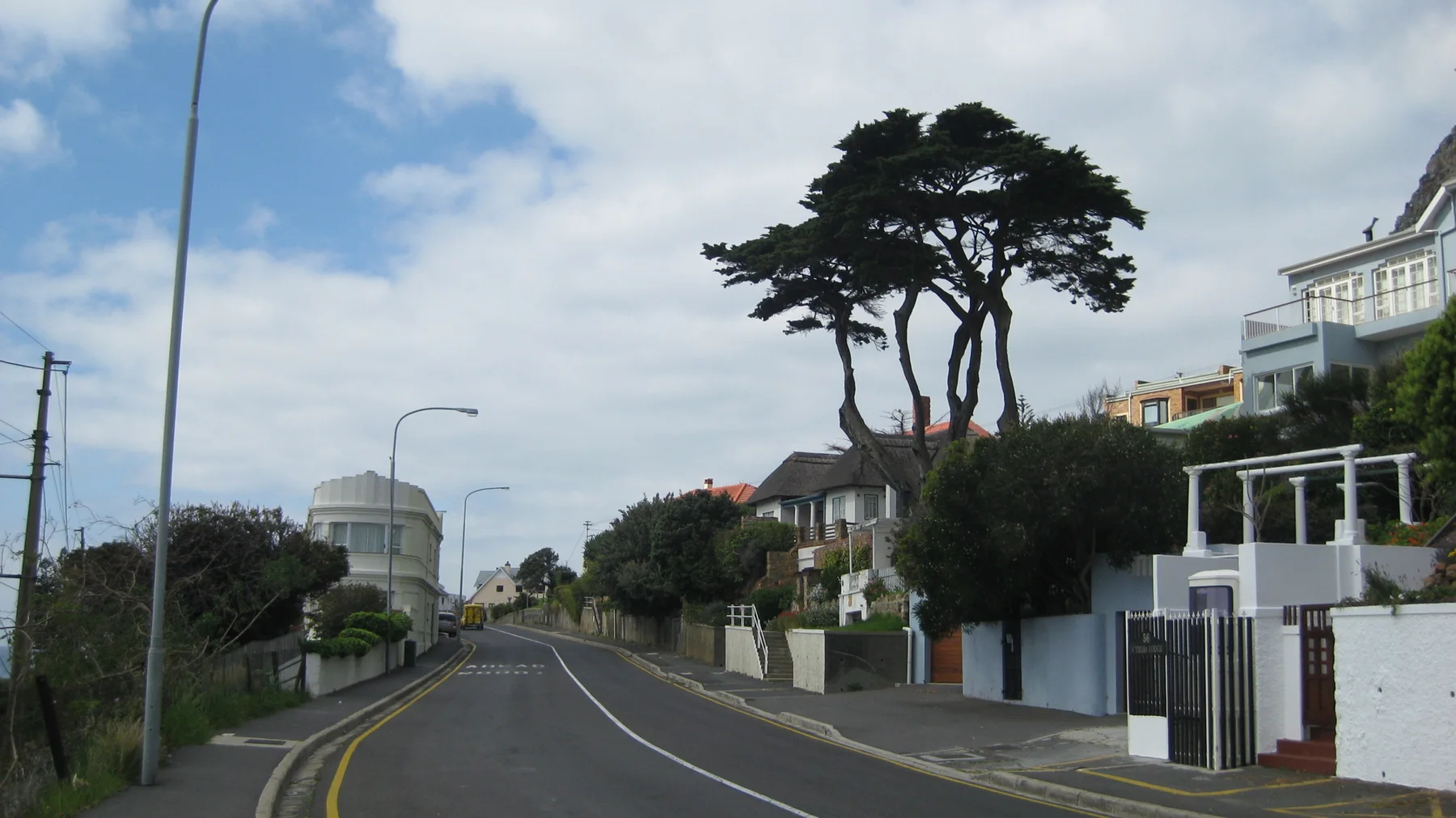 A road passes pale houses and wind-shaped trees beneath bright clouds.
