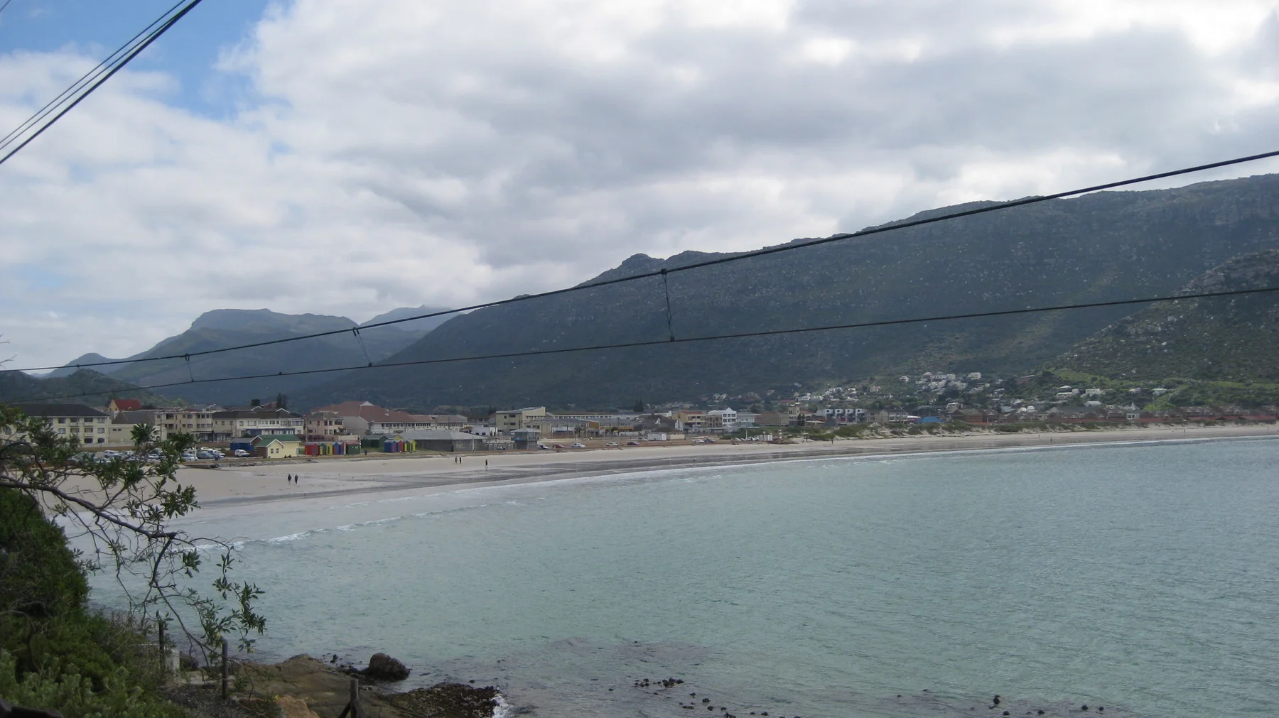 Overhead wires cross a view of coastal buildings and a broad mountain across water.