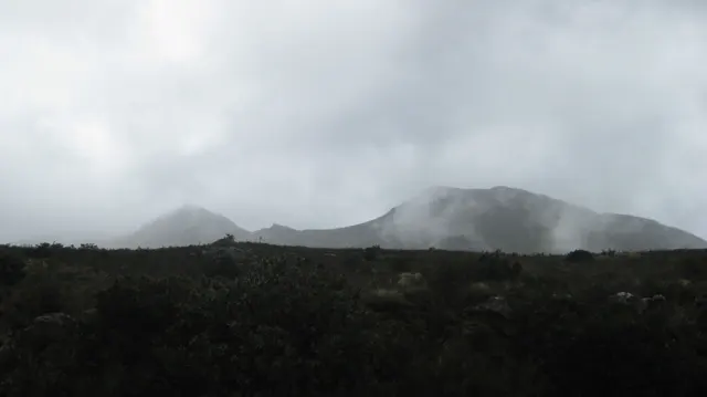 Low clouds gather around a dark mountain ridge beyond scrub.