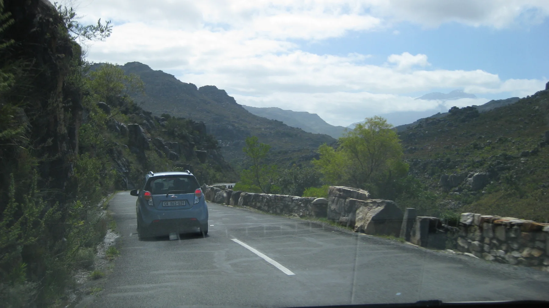 A car travels along a narrow mountain road beside a low stone wall.