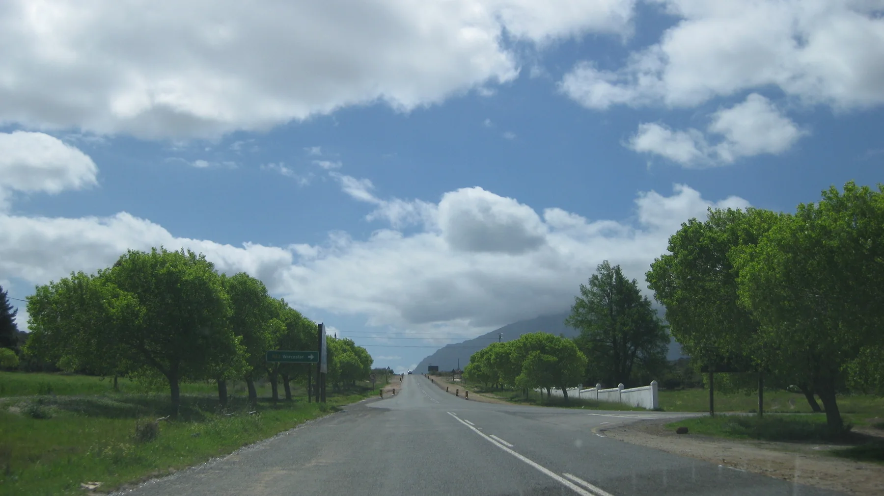A straight road runs past rounded trees beneath bright white clouds.