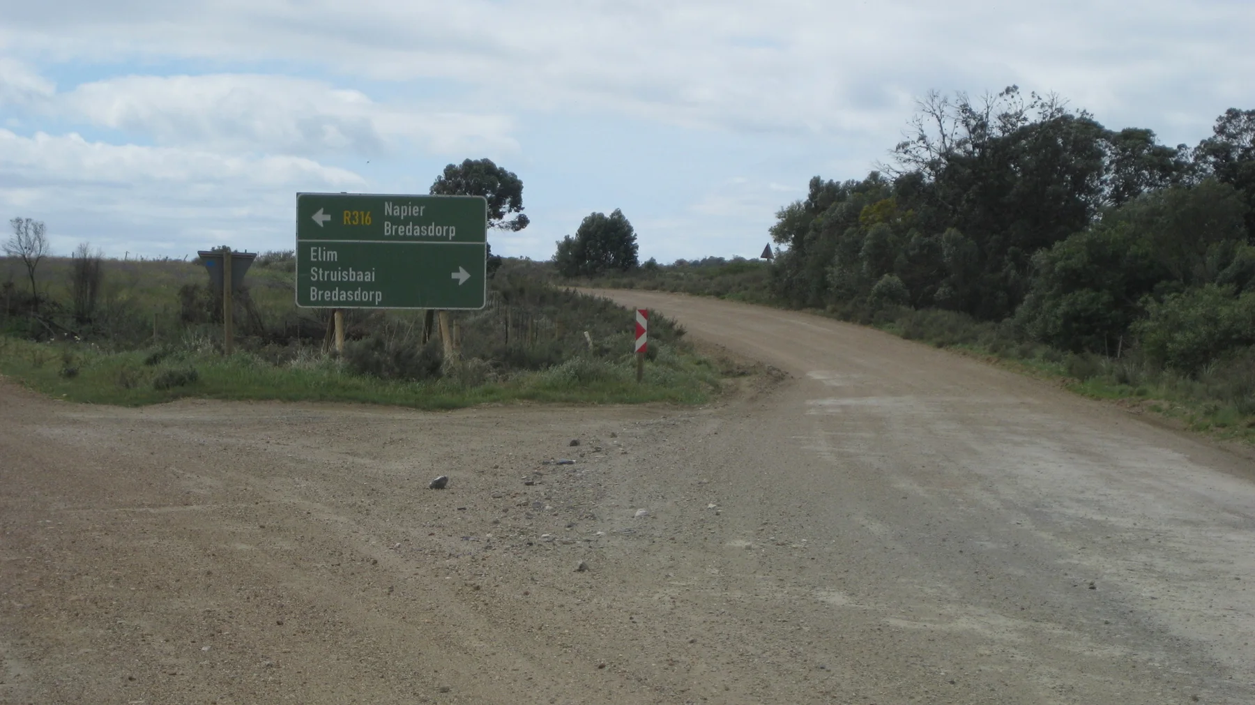 A green direction sign stands at a broad dirt-road junction.