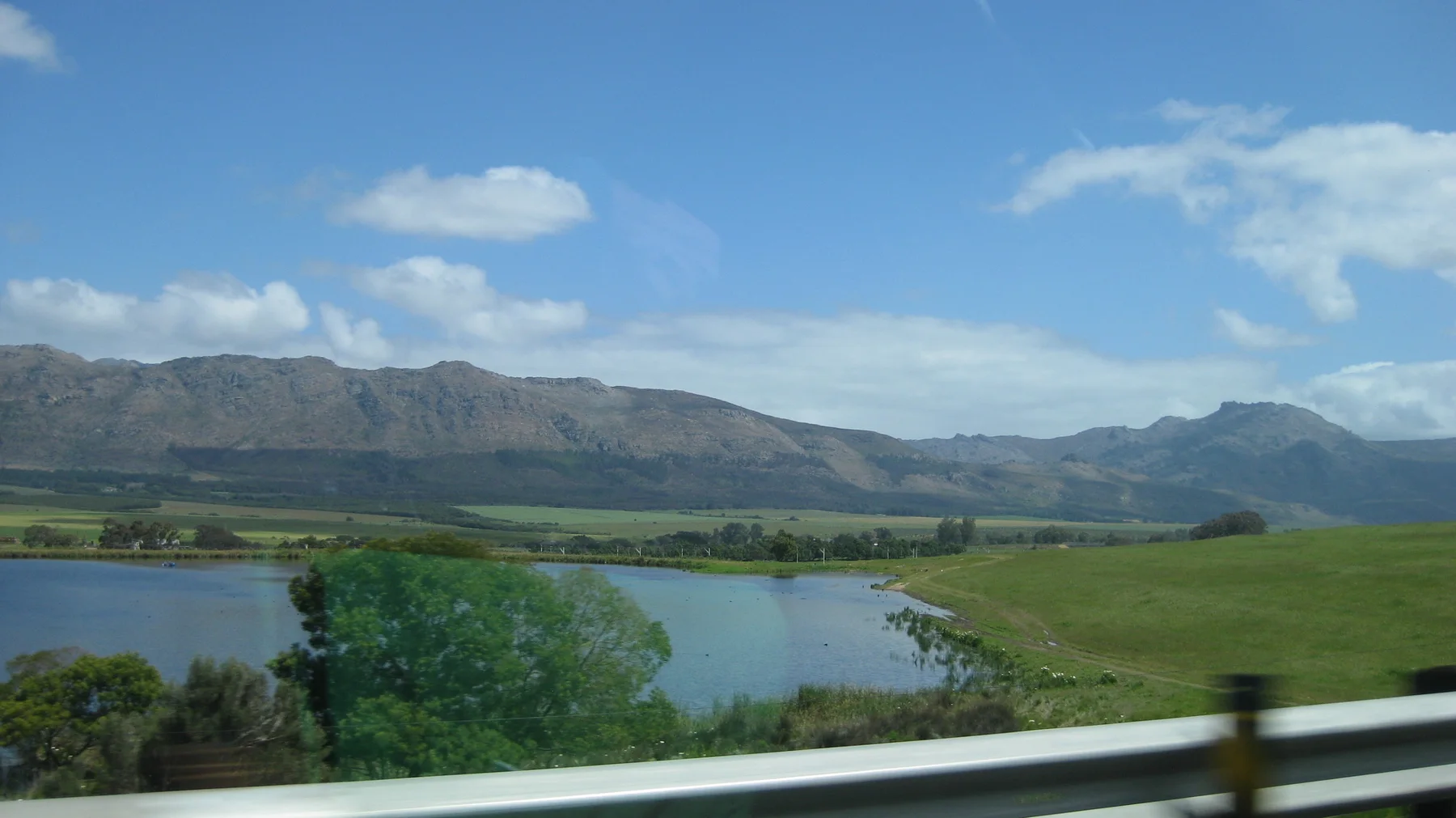 Blue water and green fields lie below a mountain range beyond a roadside barrier.
