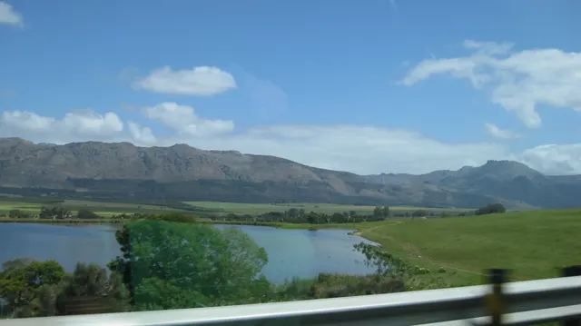 Blue water and green fields lie below a mountain range beyond a roadside barrier.
