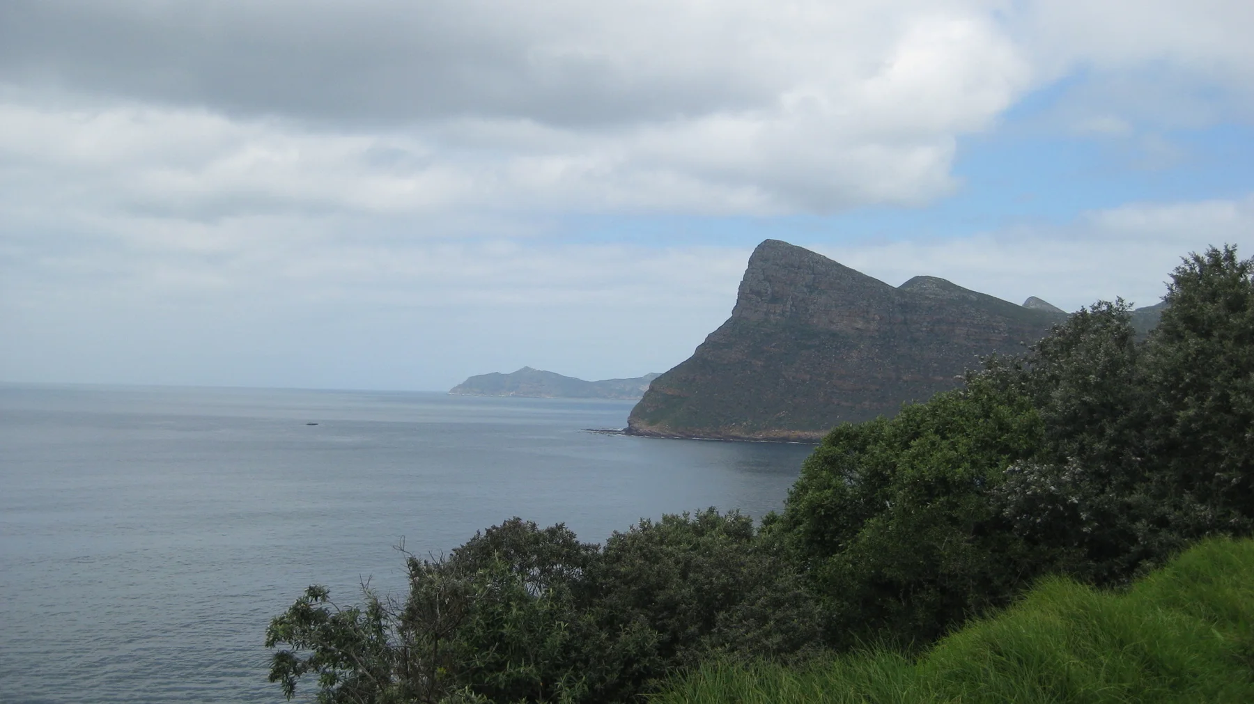 A steep headland rises beyond calm water and green coastal shrubs.