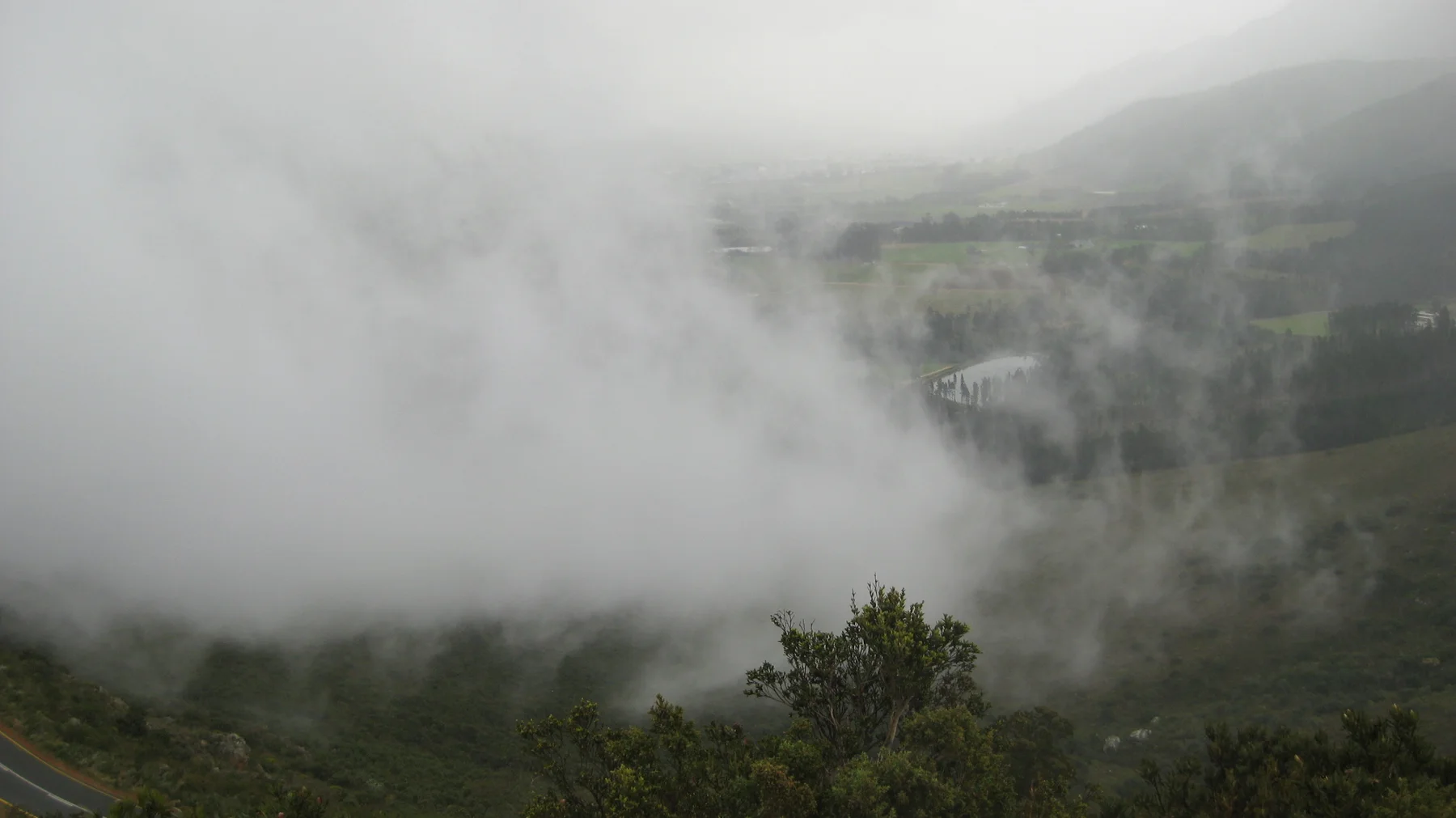 Dense mist partly hides a green valley and distant buildings.