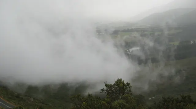 Dense mist partly hides a green valley and distant buildings.