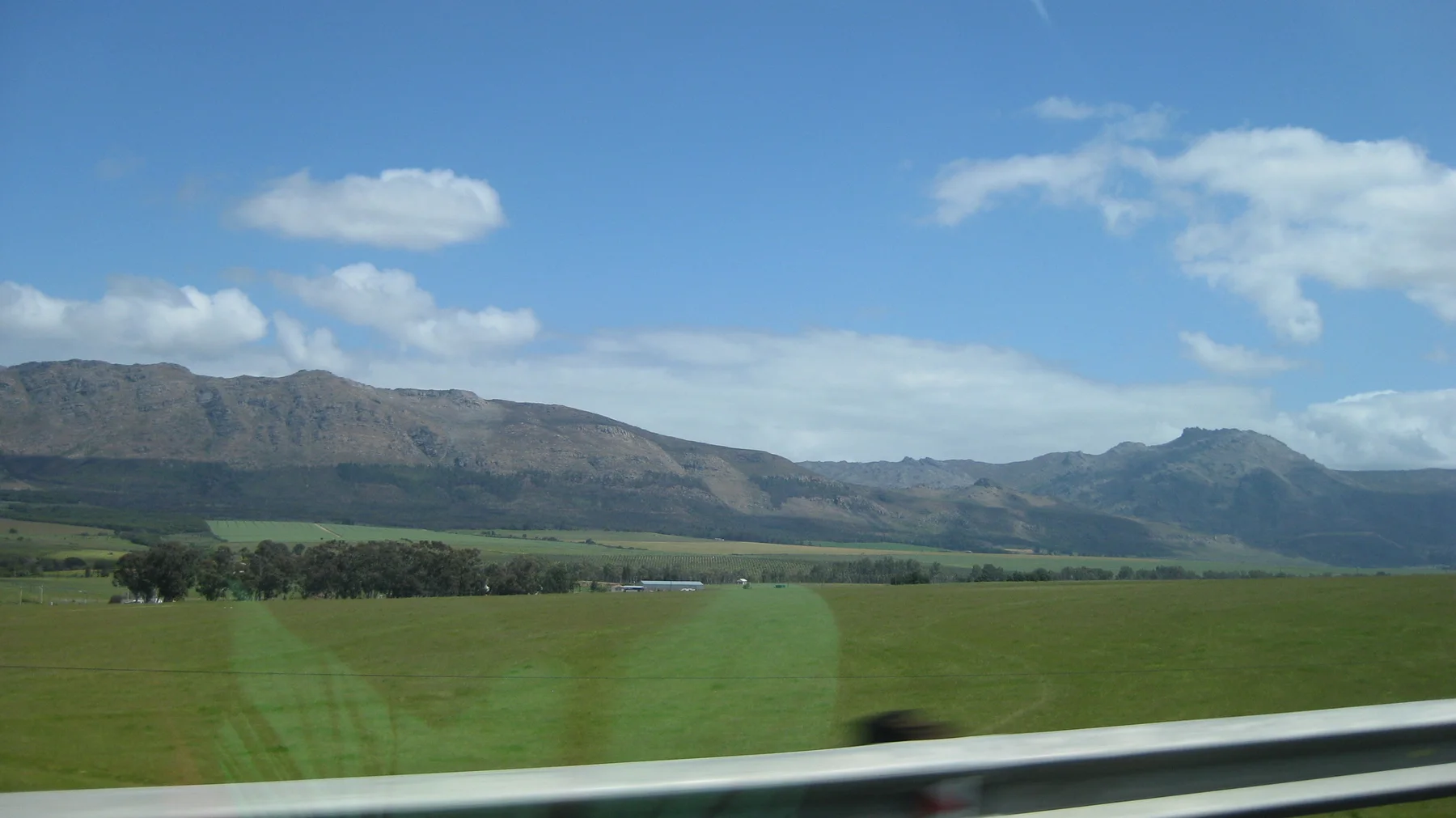 Bright green fields extend toward a mountain range beyond a metal guardrail.