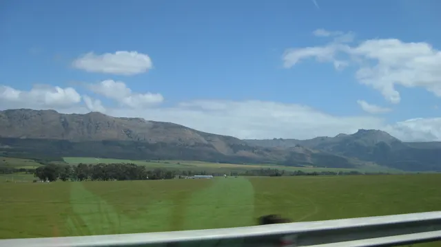 Bright green fields extend toward a mountain range beyond a metal guardrail.