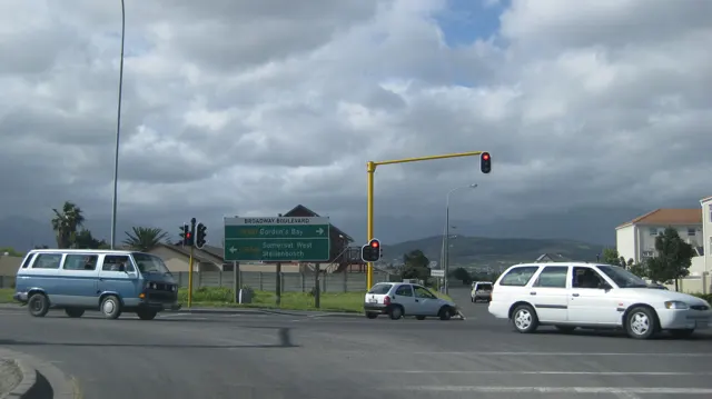 Cars wait at a junction beneath green signs and dark mountain clouds.