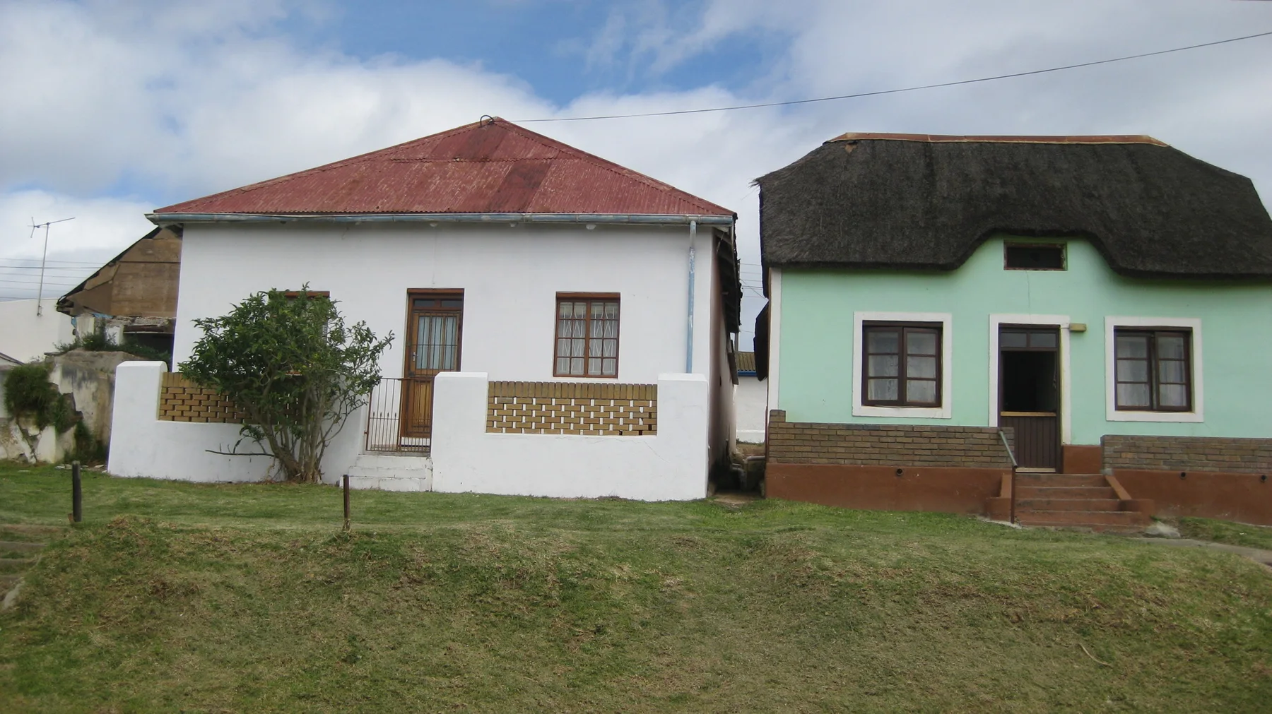 A red-roofed white house stands beside a pale green thatched house.