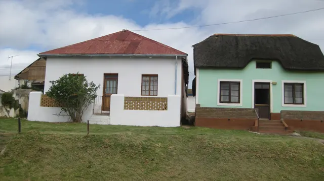 A red-roofed white house stands beside a pale green thatched house.